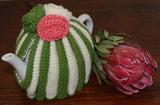 By the Tea Cosy Project - Hand knit tea cosy in protea colours, mostly green and beige striped with an added pink flower.