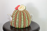 By the Tea Cosy Project - Hand knit tea cosy in a fusion of protea colours, with a green accent and flowers on top with the same colour theme.